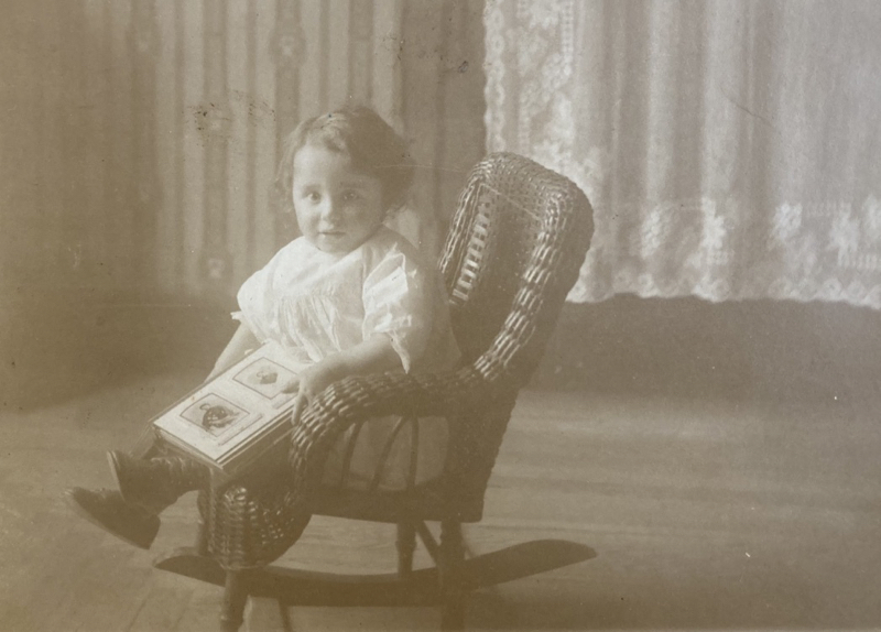Photo of Esther as a toddler in a rocking chair, probably holding a photo album